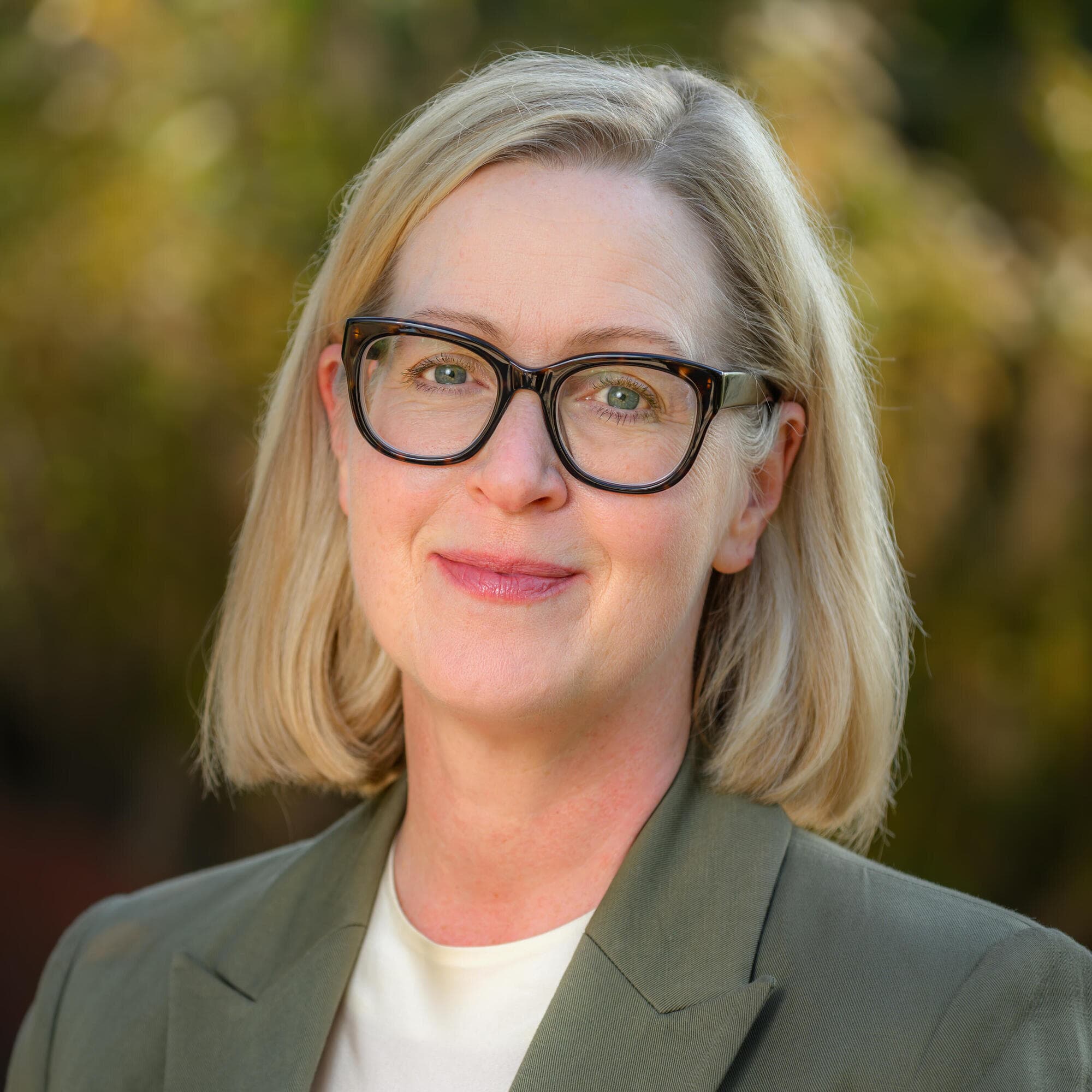 Person with glasses in an olive green blazer smiling outdoors.