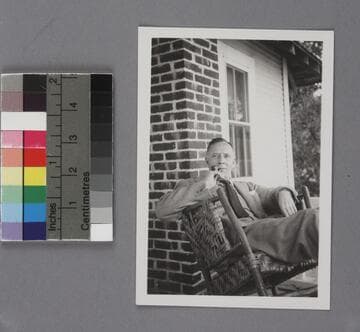 Edwin Powell Hubble seated in a woven rocking chair on a porch at Kapteyn Cottage, in front of a brick wall and clapboard wall