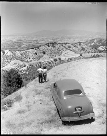 Man and woman standing by their car on a ridge overlooking valley below with mountains in background